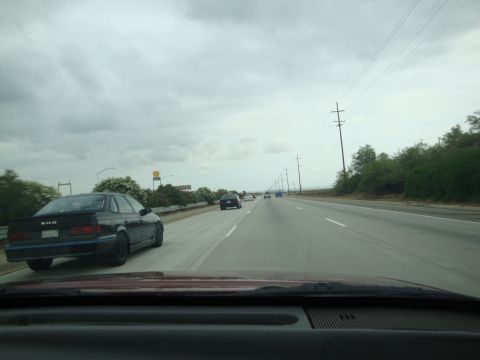 1990 Ford Taurus - A black 1990 Ford Taurus SHO sedan drives on a highway under a cloudy sky.