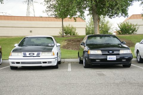 1990 Ford Taurus - Two Ford Tauruses, a white one with a black hood and a black one, are parked in a lot.