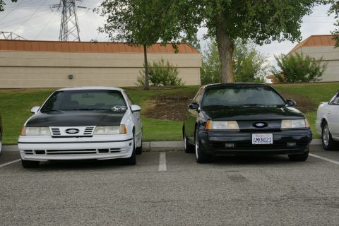 1990 Ford Taurus - Two Ford Taurus sedans are parked in a lot, one white with a black hood and the other black.