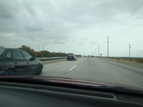 1990 Ford Taurus - Multiple vehicles travel down a highway under an overcast sky, with power lines visible along the roadside.