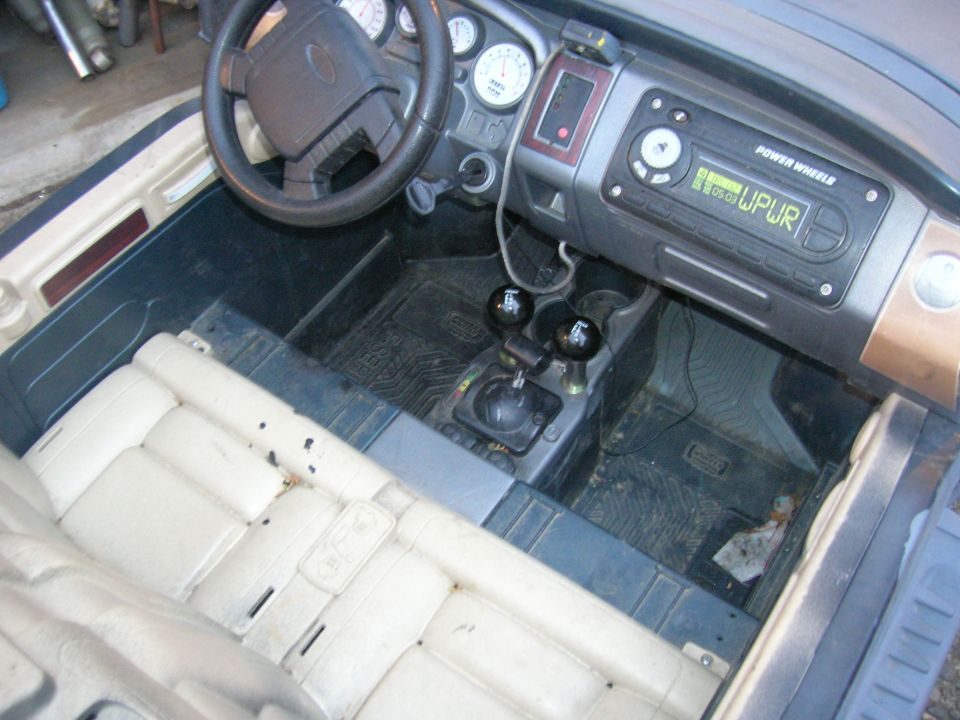 Toy car interior with beige seats, dashboard, steering wheel, and gear shift. Radio displays "POWER WHEELS UPUR"