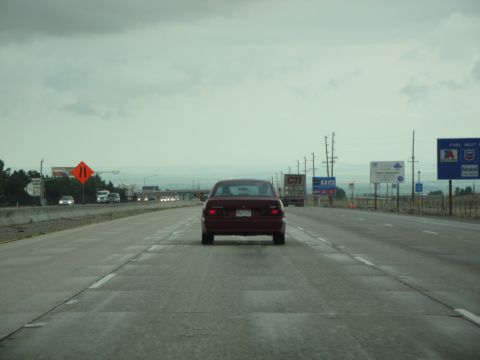 1990 Ford Taurus - A red sedan travels down a highway under an overcast sky, with other vehicles and signs visible in the distance.