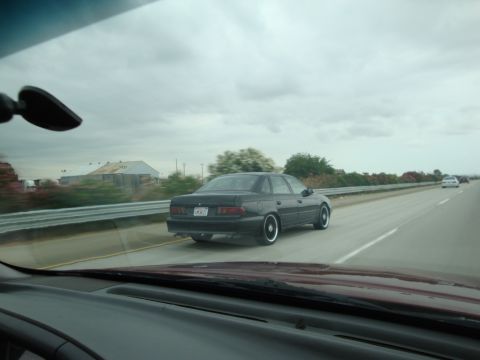 1990 Ford Taurus - A black 1990 Ford Taurus sedan is seen from behind while driving on a highway under a cloudy sky.