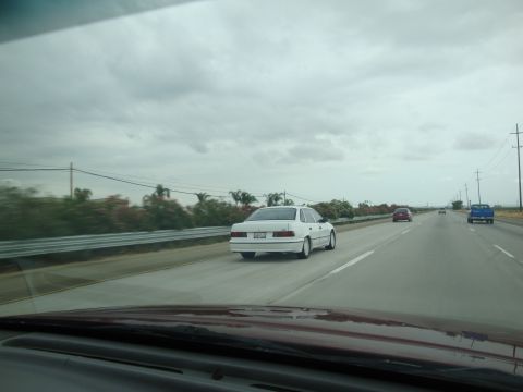 1990 Ford Taurus - A white sedan travels down the highway, its rear profile clearly visible. The vehicle appears to be from the 1990 model year.