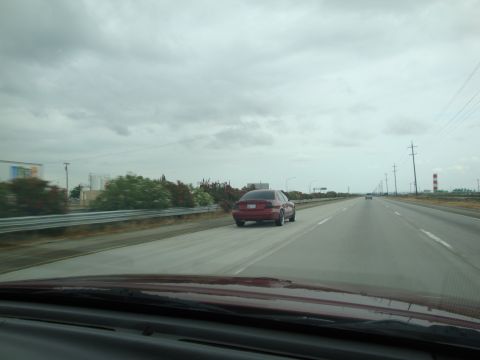 1990 Ford Taurus - A red sedan is seen from behind on a highway with industrial buildings in the distance.