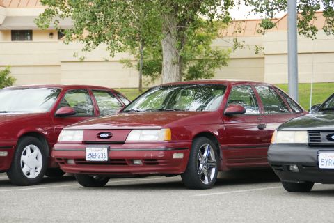 1990 Ford Taurus - A row of sedans, including a red Ford Taurus, are parked in a lot with a building and trees in the background.