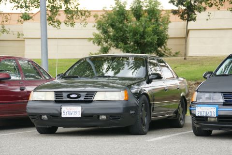 1990 Ford Taurus - A black sedan with a Ford emblem is parked in a lot, with other vehicles nearby.