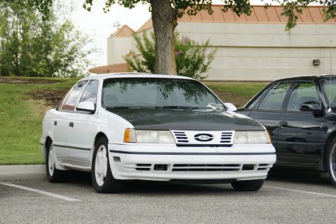 1990 Ford Taurus - A white Ford Taurus with a contrasting dark hood is parked in a lot next to another vehicle.