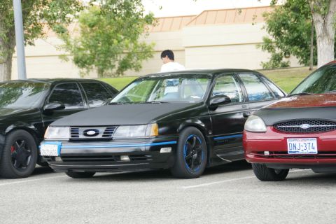 1990 Ford Taurus - A black 1990 Ford Taurus sedan with blue accents and custom wheels is parked in a lot with other cars.