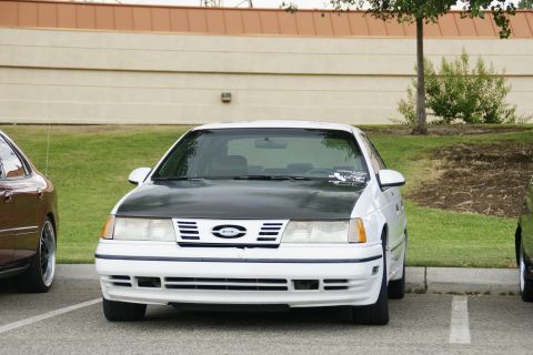 1990 Ford Taurus - A white Ford Taurus with a black hood is parked in a lot, with another vehicle visible to its left.
