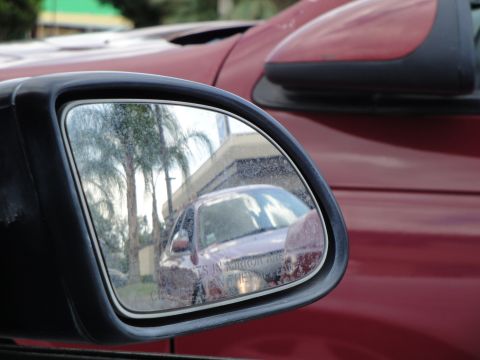 1990 Ford Taurus - Reflection in a side mirror shows a red car, palm trees, and a building. The registry claims this is a 1990 Ford Taurus, but the reflection…