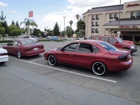 1990 Ford Taurus - A red sedan with aftermarket wheels is parked in a lot next to other vehicles.