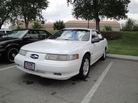 1990 Ford Taurus - A white sedan is parked in a lot next to a black car.