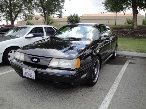 1990 Ford Taurus - A black 1990 Ford Taurus is parked in a lot, showing its front three-quarter view. Another white car is visible to its left.