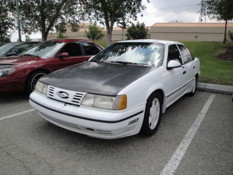 1990 Ford Taurus - A white 1990 Ford Taurus sedan is parked in a lot, with a red car beside it.