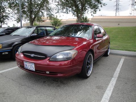 1990 Ford Taurus - A red Ford Taurus is parked in a lot next to a black car, with trees and power lines in the background.