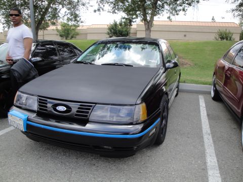 1990 Ford Taurus - A black 1990 Ford Taurus is parked in a lot, with another car to its right and a person standing to its left.
