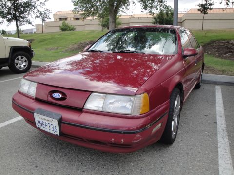 1990 Ford Taurus - A red 1990 Ford Taurus is parked in a lot, with another vehicle visible to its left.