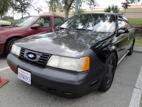 1990 Ford Taurus - A black Ford Taurus is parked next to a red car, with trees and a building in the background.