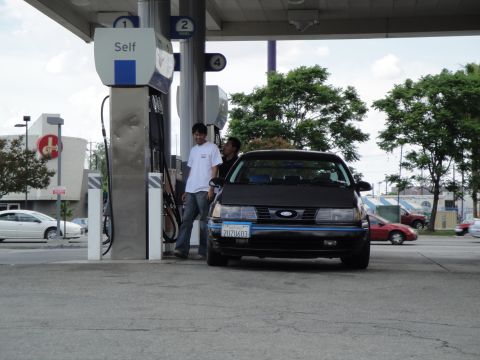 1990 Ford Taurus - A dark-colored sedan is parked at a gas station, with two individuals standing near the pump.