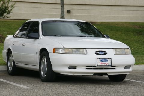 1990 Ford Taurus - This white sedan features a Ford Racing license plate, indicating a model from the mid-1990s.