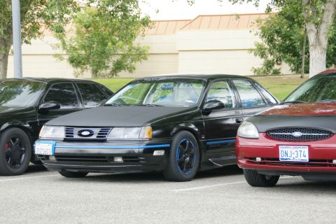 1990 Ford Taurus - Two black Ford Tauruses and a red one parked together. The black one in front has custom wheels and blue accents.