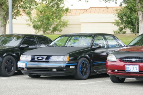 1990 Ford Taurus - A black 1990 Ford Taurus sedan with blue accents and aftermarket wheels is parked in a lot, alongside other cars.