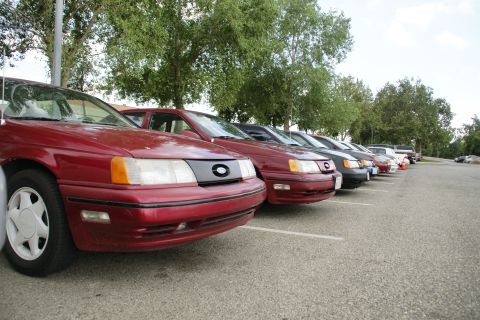 1990 Ford Taurus - Several sedans, including a red 1990 Ford Taurus, are parked in a lot on a cloudy day.