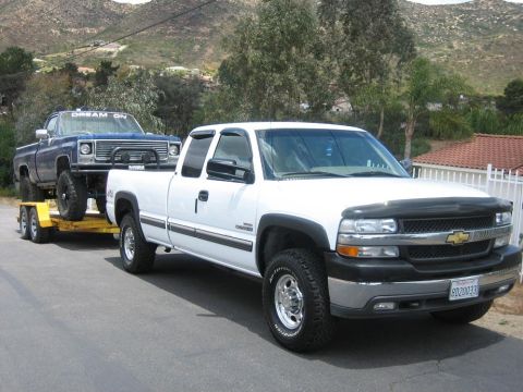 1979 Chevrolet C/K Pickup - A white pickup truck tows a blue pickup truck on a trailer down a paved road with hills in the background.