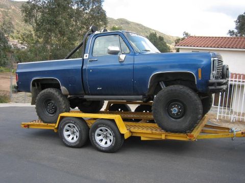 1979 Chevrolet C/K Pickup - A lifted blue pickup truck is loaded onto a yellow trailer, ready for transport.