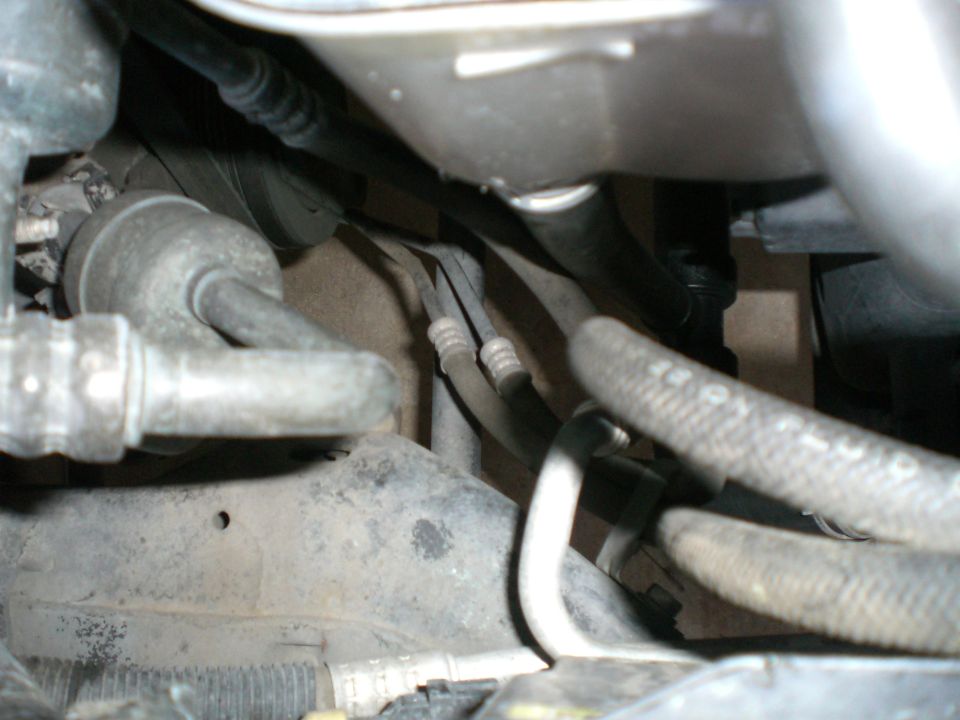 Close-up of dirty engine bay hoses, hard lines, and components under a metal reservoir