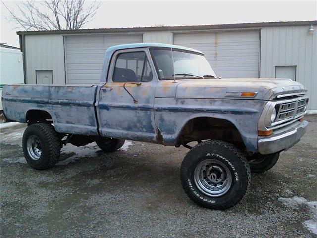 Faded blue-gray Ford F-100 pickup, lifted on off-road tires, parked outside a metal building
