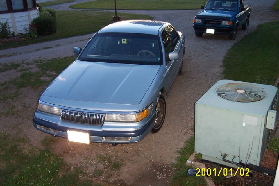 Light blue Mercury Grand Marquis sedan parked on gravel driveway near an AC unit, pickup truck behind