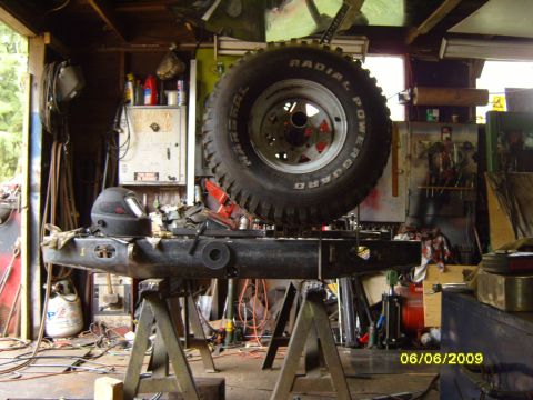 1989 Ford Bronco - A black steel rear bumper is supported by sawhorses, with a spare tire suspended overhead in a workshop.