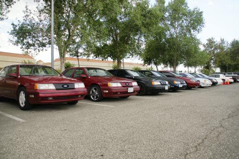 1990 Ford Taurus - A row of sedans and a coupe are parked in a lot on a cloudy day.