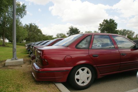 1990 Ford Taurus - A row of cars are parked in a lot, with the side of a red 1990 Ford Taurus in the foreground.