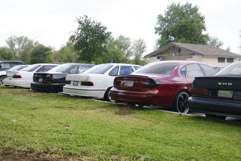 1990 Ford Taurus - A line of sedans, including several Ford Taurus models, are parked in a lot on a cloudy day.