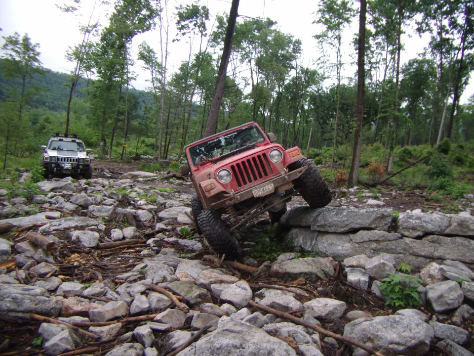 Red Jeep Wrangler climbing over a rock obstacle on a wooded trail; white Hummer in background