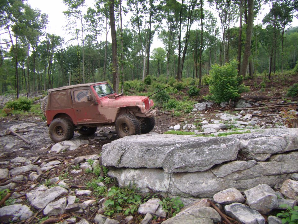 Red Jeep Wrangler driving over rocks on a wooded trail. It's covered in mud