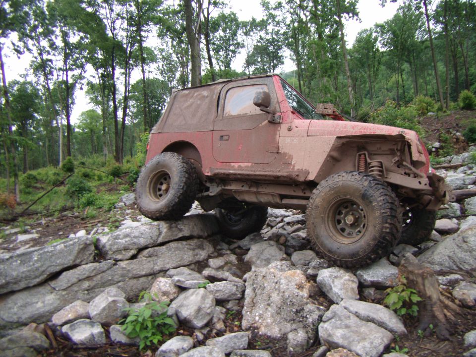 Red Jeep Wrangler with soft top navigating a rocky trail, covered in mud