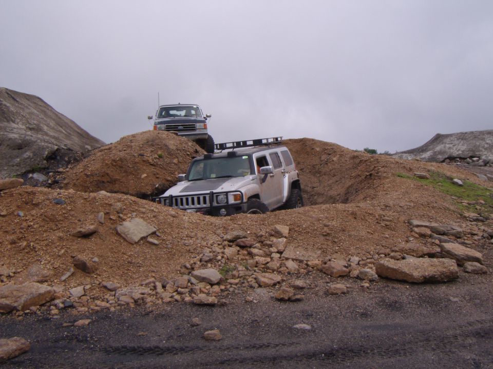 Silver Hummer H3 with roof rack stuck in dirt pit, pickup truck on hill behind