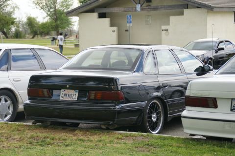 1990 Ford Taurus - A black 1990 Ford Taurus is parked next to other vehicles, showing its rear end and aftermarket wheels.