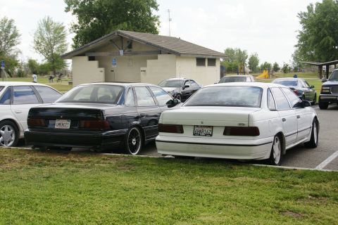 1990 Ford Taurus - Two Ford Taurus sedans, a black and a white model, are parked in a lot with a building in the background.