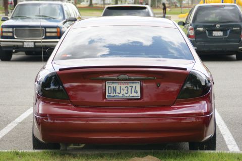 1990 Ford Taurus - Rear view of a red sedan with tinted taillights and a spoiler. Other vehicles are parked nearby.