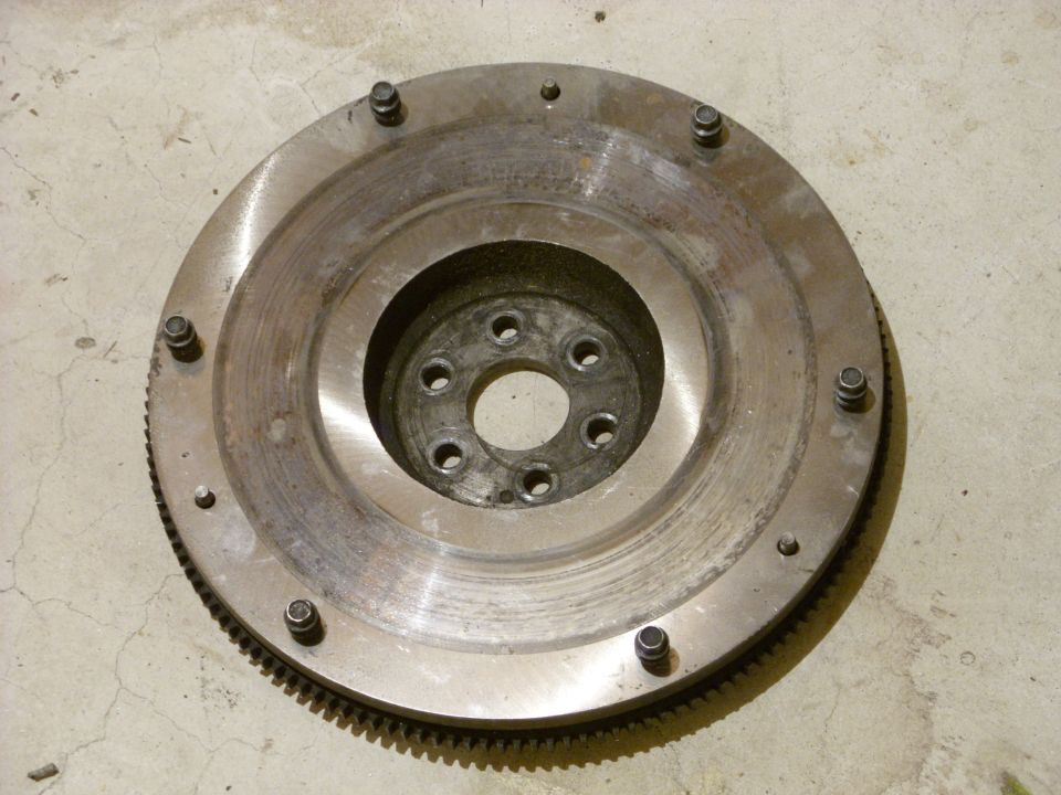 Metal flywheel with bolt holes and gear teeth around the edge, sitting on a concrete floor