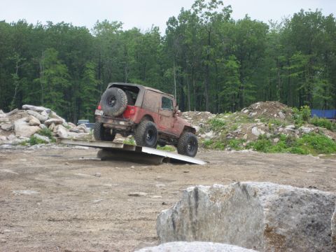 1991 Ford Bronco - A red Jeep Wrangler TJ navigates an off-road obstacle course with a wooded area behind it.