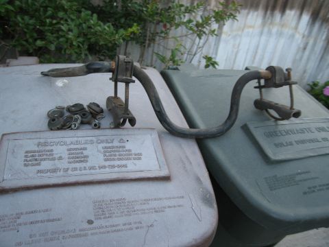 1986 Ford Bronco - A metal part, likely for a vehicle, is displayed with its associated hardware on top of two recycling bins.