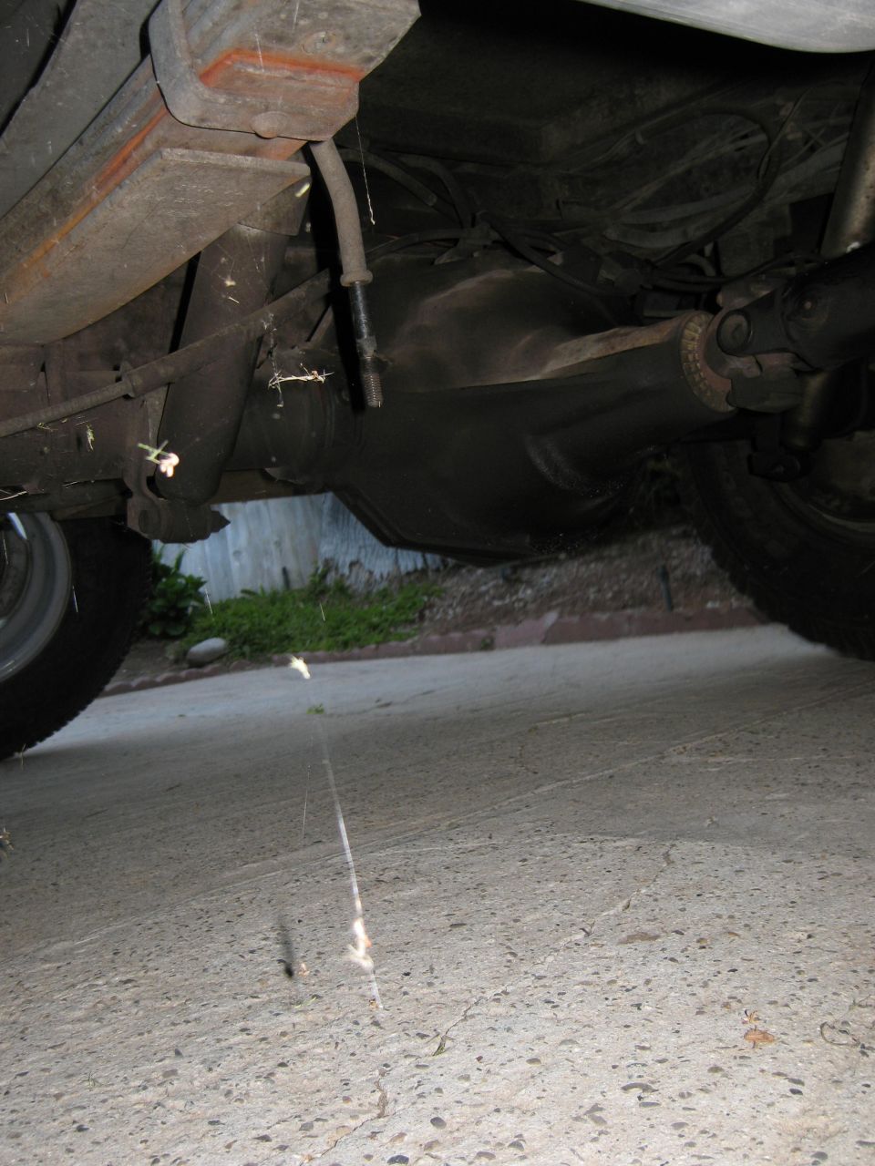 Underside of a vehicle showing the rear axle, leaf springs, and driveshaft. A tire is visible on the left