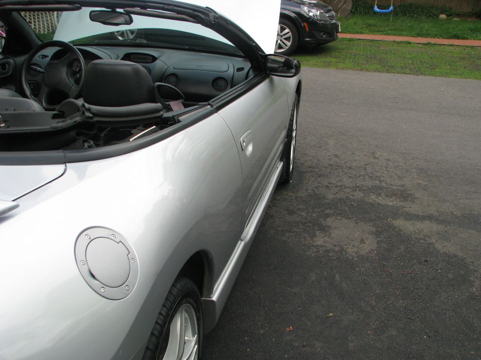 Silver convertible sports car, driver's side view, showing door handle, gas cap, and interior. Another car is parked behind it
