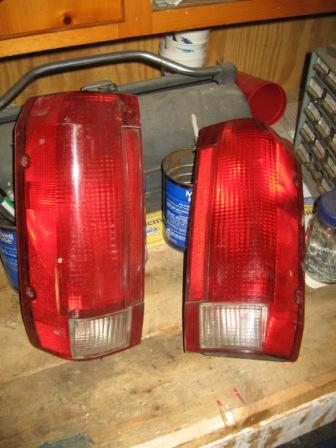 1993 Ford Bronco - A pair of red Ford truck taillight assemblies with clear backup-lamp lenses at the bottom, standing upright on a cluttered wooden workbench…
